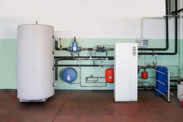 A modern heating system with a large water tank, a white control unit, red expansion vessels, various pipes, valves, and a blue heat exchanger against a green and white wall.